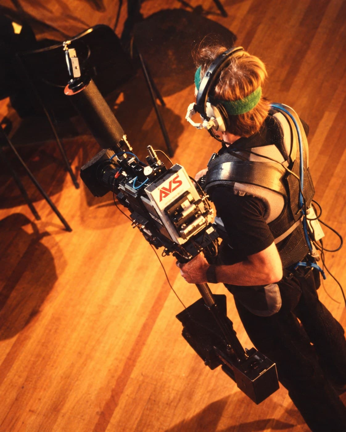 Steadicam operator in a headset and vest carrying a live-TV Steadicam rig across a wooden stage, seen from above
